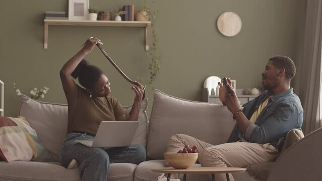 Medium slowmo of young Black man taking photo on smartphone of his girlfriend posing with pet snake in hands sitting on sofa in minimalist eco style living room with olive green walls and houseplants