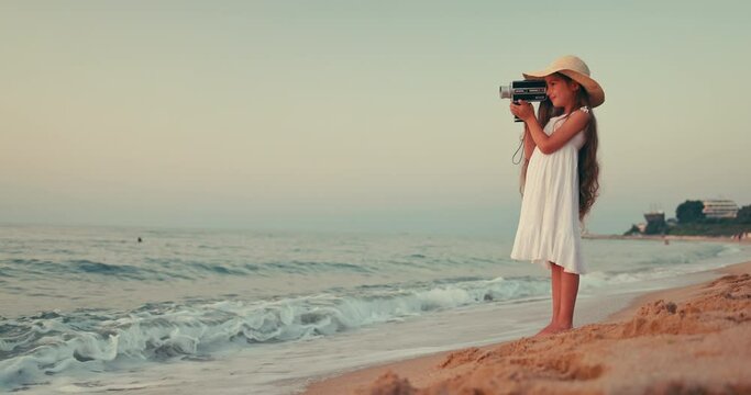 Girl with white dress and long hair making pictures and video with old vintage camera of a sunrise on the beach