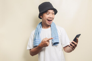 asian man wearing bright white t-shirt in black hat with blue towel shocked to see his phone on isolated background