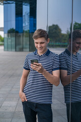 Young caucasian man in casual clothing standing near modern glass building and looking at his phone. People using phones. High quality photo