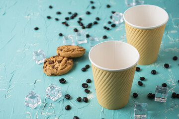 Top view of two cardboard cups on a blue table with ice, cookies and coffee beans, horizontally, with copy space