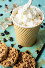 Aerial view of a cardboard cup with coffee and cream on a blue table with spilled coffee, ice, cookies and coffee beans, vertical