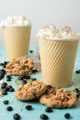 Close-up of two cardboard cups with coffee and cream on a blue table with spilled coffee, cookies and coffee beans, vertical