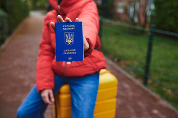 The girl sits on a suitcase and keeps a close-up passport of Ukraine. Refugees and IDPs from...