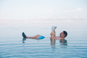 Man lies in blue water of Dead Sea. Person with newspaper in hands. Very salty water push out. Treatment and recreation on beach, travel and holidays in Israel. Salt and mud healthy for body