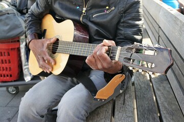 Mann mit schwarzer Lederjacke und grauer Jeanshose sitzt auf Bank und spielt Gitarre in Stadt 