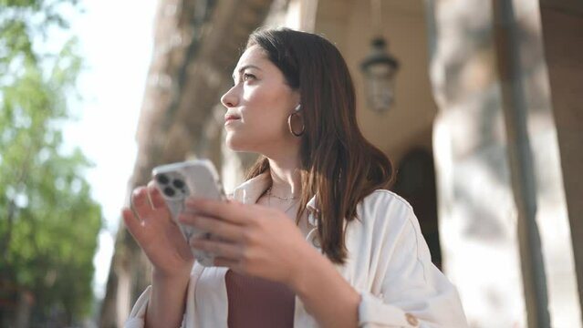 Pensive brunette woman in headphones looking around and texting on phone on the street