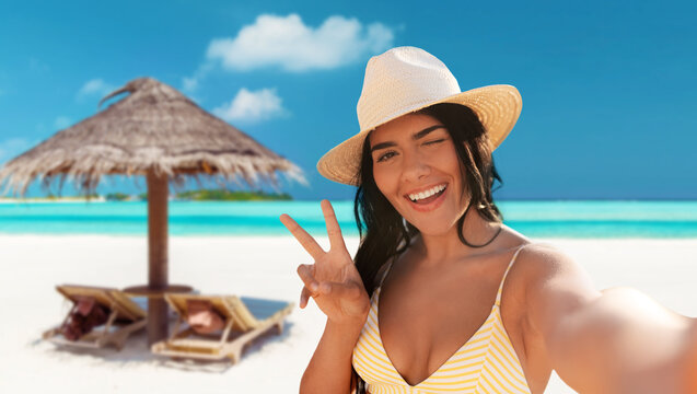 people, summer and swimwear concept - happy smiling woman in bikini and straw hat taking selfie and showing peace gesture over palapa and sun beds on tropical beach background in french polynesia