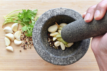 Garlic, peppercorns and coriander roots in a gray granite mortar.  Hand grinding garlic, mixed peppercorns and coriander roots in a mortar and pestle. Cooking preparation concept. 