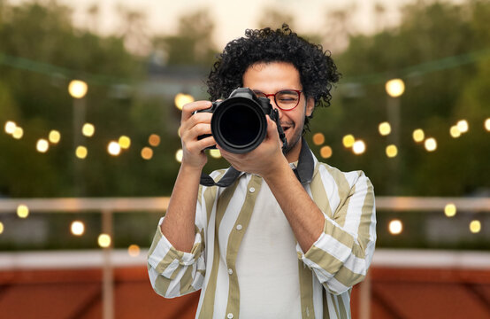 photography, profession and people and concept - happy smiling man or photographer in glasses with digital camera over party lights on roof top background