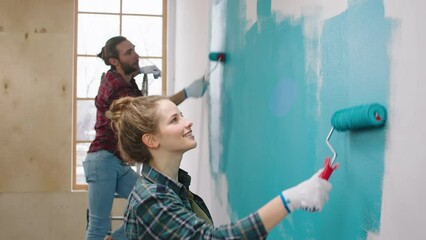 At home charismatic woman and her husband Caucasian looking painting walls in a blue colour smiling large and feeling happy working together - Powered by Adobe