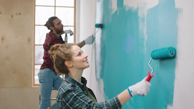 At home charismatic woman and her husband Caucasian looking painting walls in a blue colour smiling large and feeling happy working together