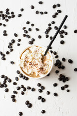 Aerial view of cardboard cup with coffee with cream and black straw, on white table with coffee beans, selective focus, vertical, with copy space
