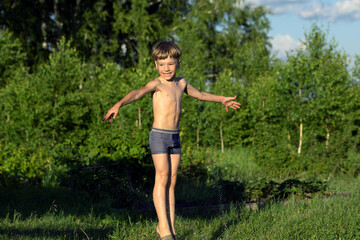 Happy smiling boy in wet shorts plays with water sprinkler on natural blurred background. Summer games on vacation. Happy life.