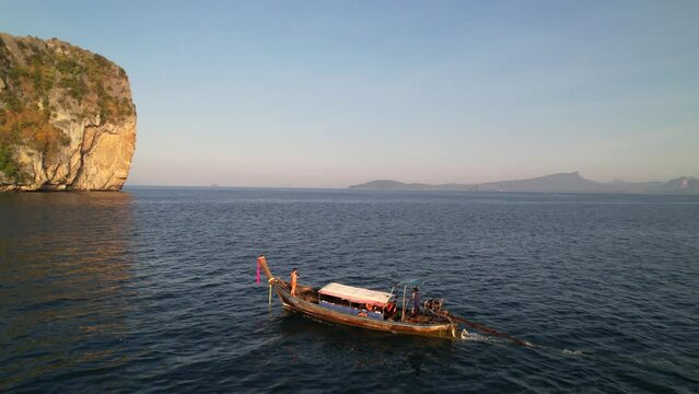 aerial drone approaching a thail longtail boat anchored in the water of Krabi Thailand during a sunrise tour with a tourist on board and a large limestone rock in the distance