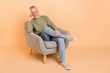 Full length photo of funky elder grey hairdo man sit wear eyewear pullover jeans shoes isolated on beige background