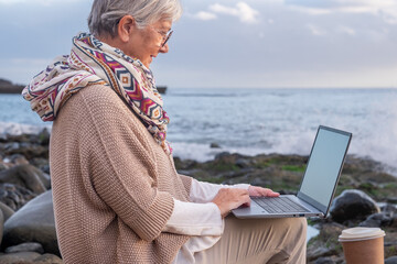 Senior active woman sitting on the beach using laptop wearing scarf. Elderly attractive female in remote work enjoying sunset at sea, horizon over water