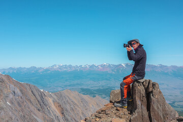 Tourist sitting on stone near abyss edge on high altitude under blue sky in sunny day. Man with camera on high rock near precipice edge with wonderful view from above to large mountain range in away.