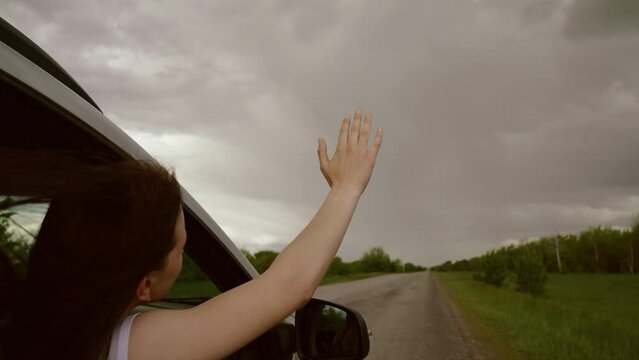 Road trip on way to vacation, Free girl waves her hand from car window, travels, catches wind with her fingers, Clouds. Young female driver plays, catches fresh wind with her hand from car window