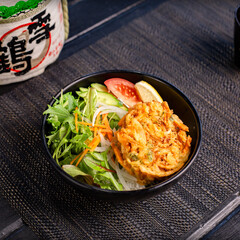 Veggie vegan tempura don with salad in the traditional Japanese ramen restaurant, with a black bowl on a black mat and Japanese fonts sake barrel translation: Snow Crane