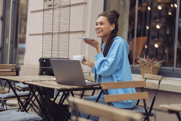 Pretty cheerful woman sending voice message and having coffee at cafe terrace. Audio chat. Outside.