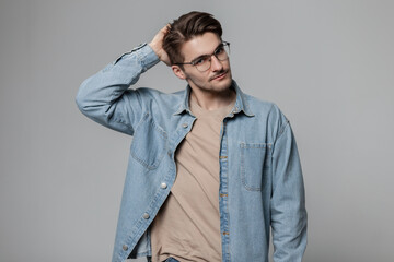 Fototapeta premium Cool handsome fashion student man with hairstyle in blu jeans shirt with vintage stylish glasses stands and poses on a gray background in studio