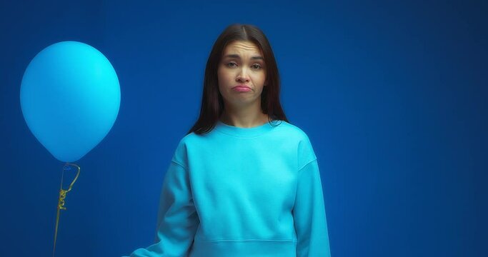 An upset oriental-looking woman wearing a blue jumper shows a sad and unhappy face because of one balloon on an isolated blue background