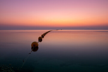 Seascape showing a pink sunrise in dim light over the calm waters of the Mar Menor, in the Region of Murcia, Spain. On the water, a line of yellow buoys that are lost in the horizon.