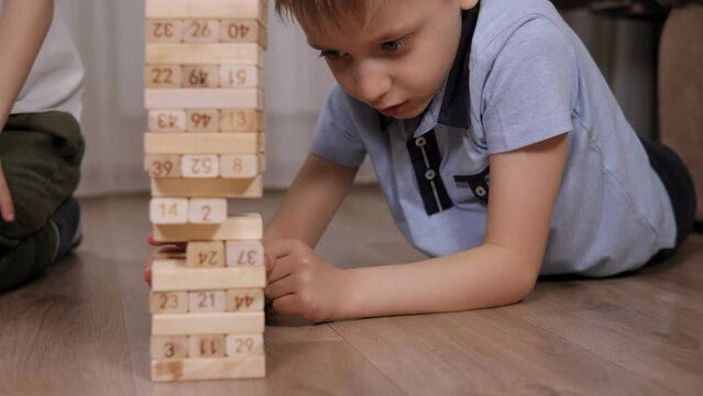 Close-up of two positive boys playing a fun active game of wooden blocks at home, the child takes out one wooden block and the tower falls.