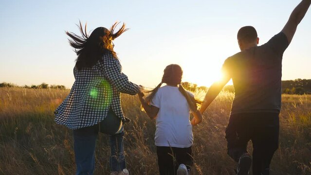 Mother and father with small daughter jogging with multicolored kite through grass field at sunset. Young parents with little cute girl holds hands of each other running among summer meadow. Rear view
