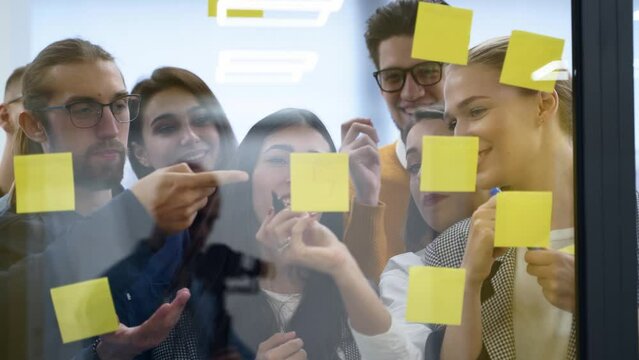 Business team brainstorm writing ideas on paper sticky notes and post it on glass wall. Startup coworkers discuss a strategy of work plan for product development at scrum meeting in corporate office.