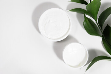 A mock up white jar of cream on a white gray background, on a white table with green leaves of a tropical plant, with hard shadows. Stylish look of the product, identity.