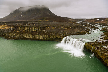 Greenish water stream falls over lava cliff into green lake, background brown Icelandic peak covered with cloud layer and grey sky