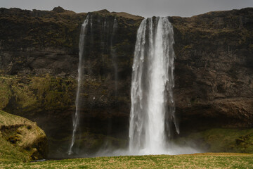Shiny white waterfall frontal in center from bottom over dark cliff, foreground green-yellow meadow, sky monotonous grey