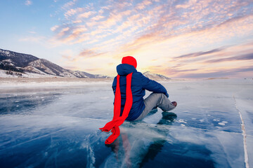 Travel winter Lake Baikal, smile man tourist in red cap sits on ice sunset