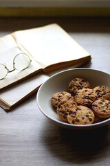 Plate of chocolate chip cookies, open books and reading glasses on wooden table. Selective focus.