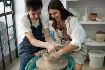 Man with young woman works on potter's wheel.  lovely couple have fun, sculpt pot from white clay.  Man hugs woman.  Studio on production of clay tableware love and tender.