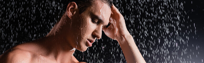 man with closed eyes touching hair while showering on black background, banner.