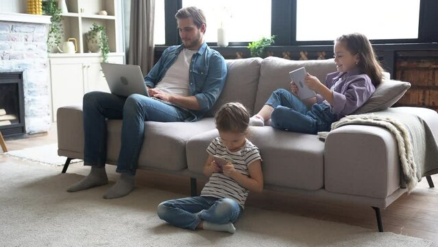 Addicted to gadgets father and kids busy on laptop, tablet and cellphone while sitting on sofa in living room. Modern family with children spending joint leisure time using gadgets