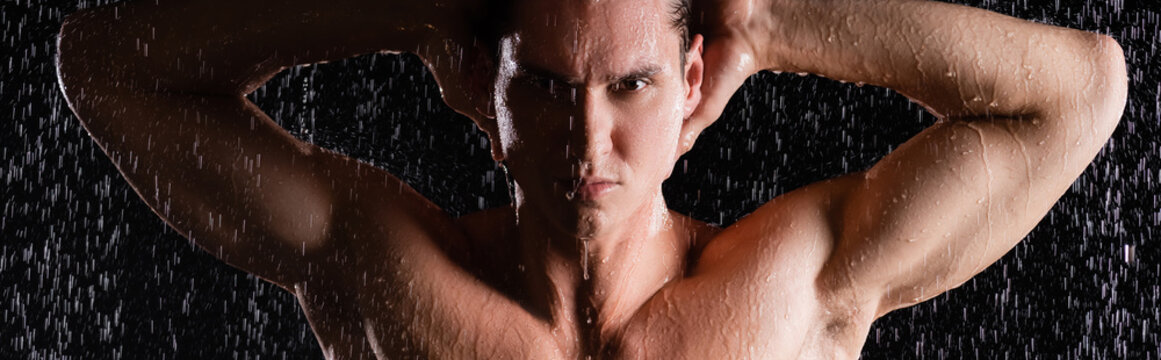 muscular man posing with hands behind head under shower on black background, banner.