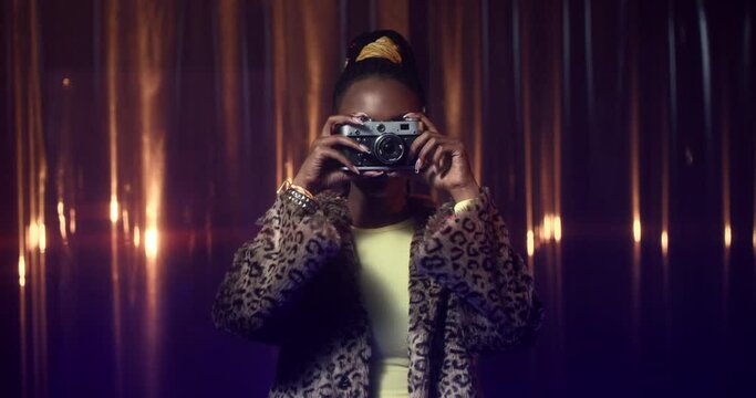 Young African American woman in fur coat and skinny top makes photos with film camera, then smiles. Happy girl in party outfit with bold makeup taking pictures.