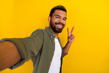 Photo of excited funky guy dressed green shirt tacking selfie showing v-sign isolated yellow color...