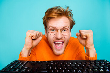 Photo of shocked lucky guy dressed orange sweater chatting keyboard rising fists isolated turquoise...