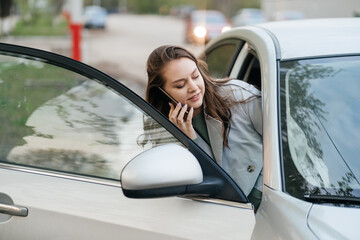 Naklejka premium beautiful girl with long hair in a grey trench coat using smartphone call gets into the car
