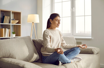 Obraz premium Calm young Caucasian woman sit on couch with mudra hands practice yoga meditate. Relaxed girl have meditation session on sofa at home, relieve negative emotions breathe. Stress free concept.