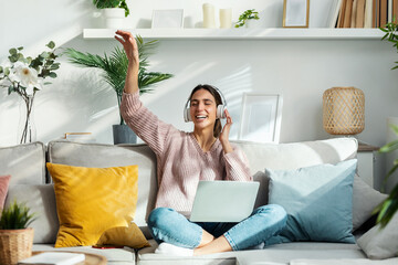 Motivated young woman listening to music with laptop while singing and dancing sitting on sofa at...