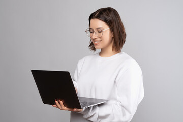 Concentrated smiling woman wearing glasses is holding a laptop while standing. Studio shot over grey background.