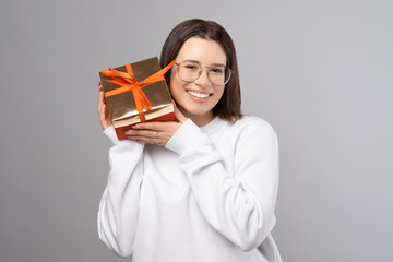 Joyful woman wearing glasses is holding a red gift box near her face. Studio shot over grey background. © Vulp