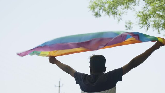back view shot of young man proudly holding waving lgbtq flag - cocnept of pride month celebration, support and movement.
