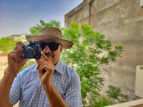 Picture of a person wearing sunglasses and hat out in a sanctuary in summers with green leaves in background shooting pictures with his camera